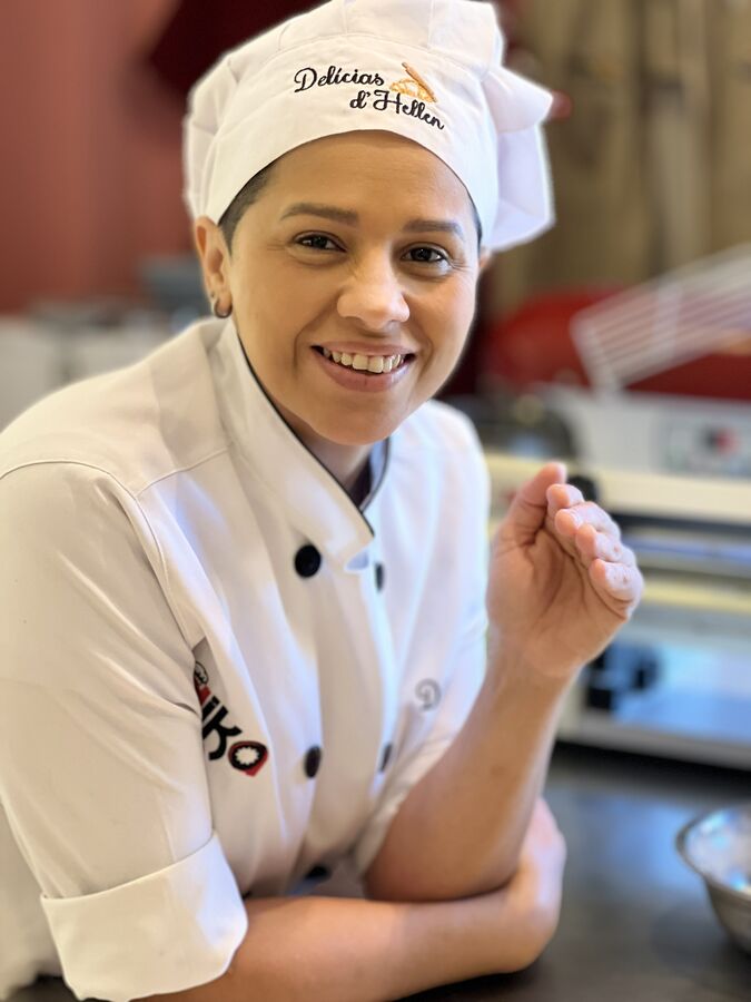 Hellen Miranda, de dólmã e touca bordada Delícias d'Hellen, sorrindo na cozinha de produção